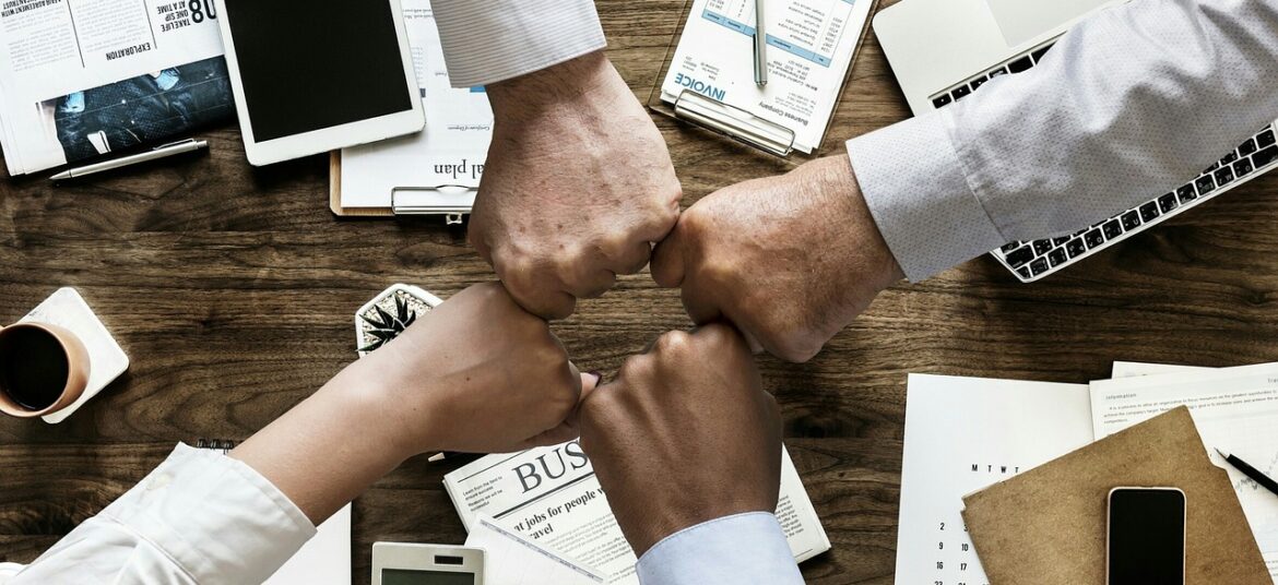 Vier Geschäftspersonen vereinen ihre Fäuste über einem Schreibtisch – Symbol für Teamarbeit, flexible Recruiting-Strategien, krisenfestes Recruiting, externe Personalgewinnung.